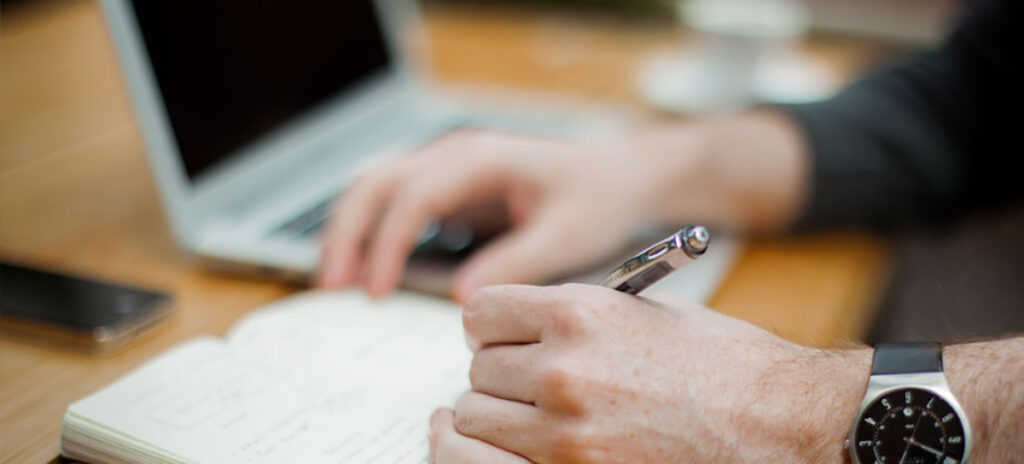 A person writing in a notebook next to a laptop