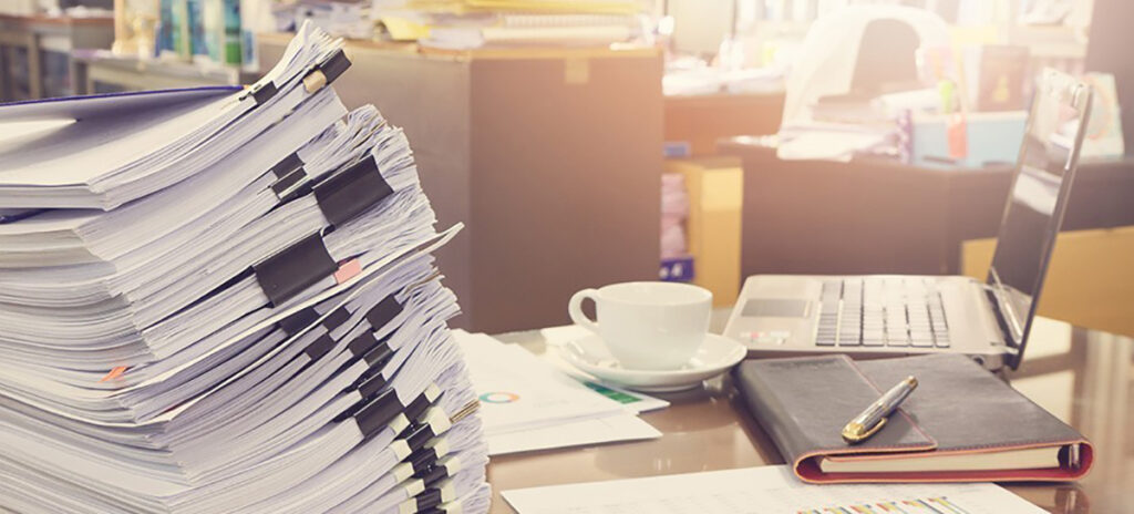 A cluttered desk with piles of paper, notebooks, a laptop and coffee