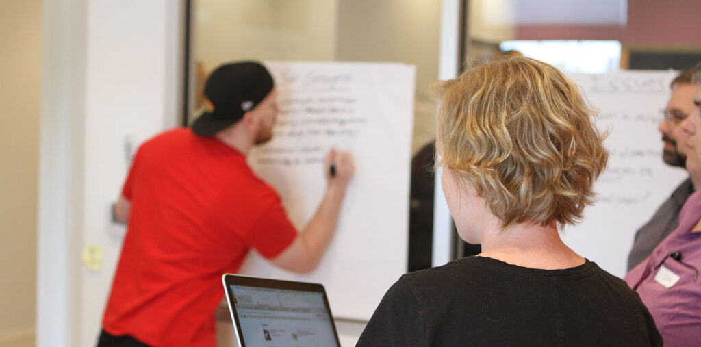 Photo from a Project Context Workshop, person writing on a poster on the wall while team member holds a laptop for reference