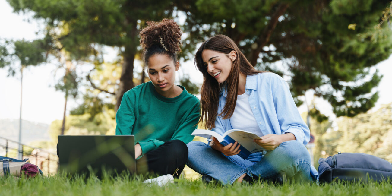 Two female university students sitting in the grass under a tree looking at a laptop