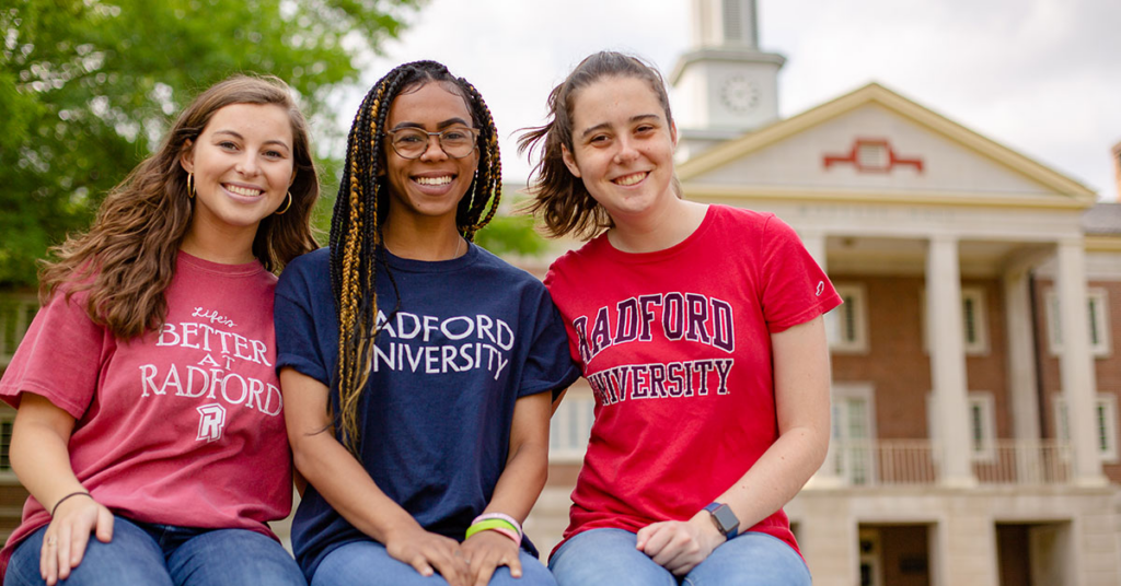 Three female students wearing different colored Radford University t-shirts sit smiling in front of one of the campus buildings