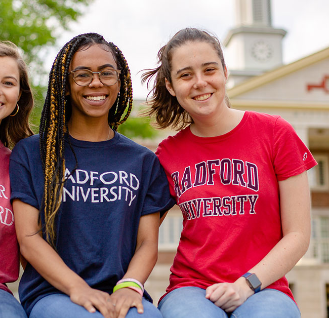Three female students wearing different colored Radford University t-shirts sit smiling in front of one of the campus buildings
